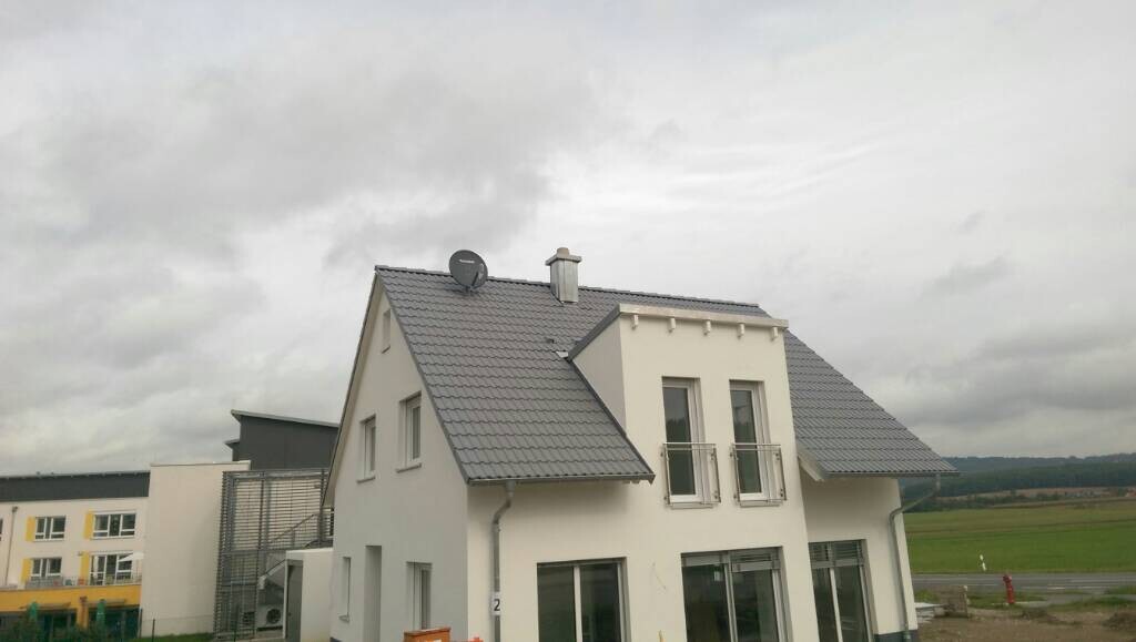 Modernes weißes Einfamilienhaus mit grauem Satteldach vor grünem Feld und Wolkenhimmel.
