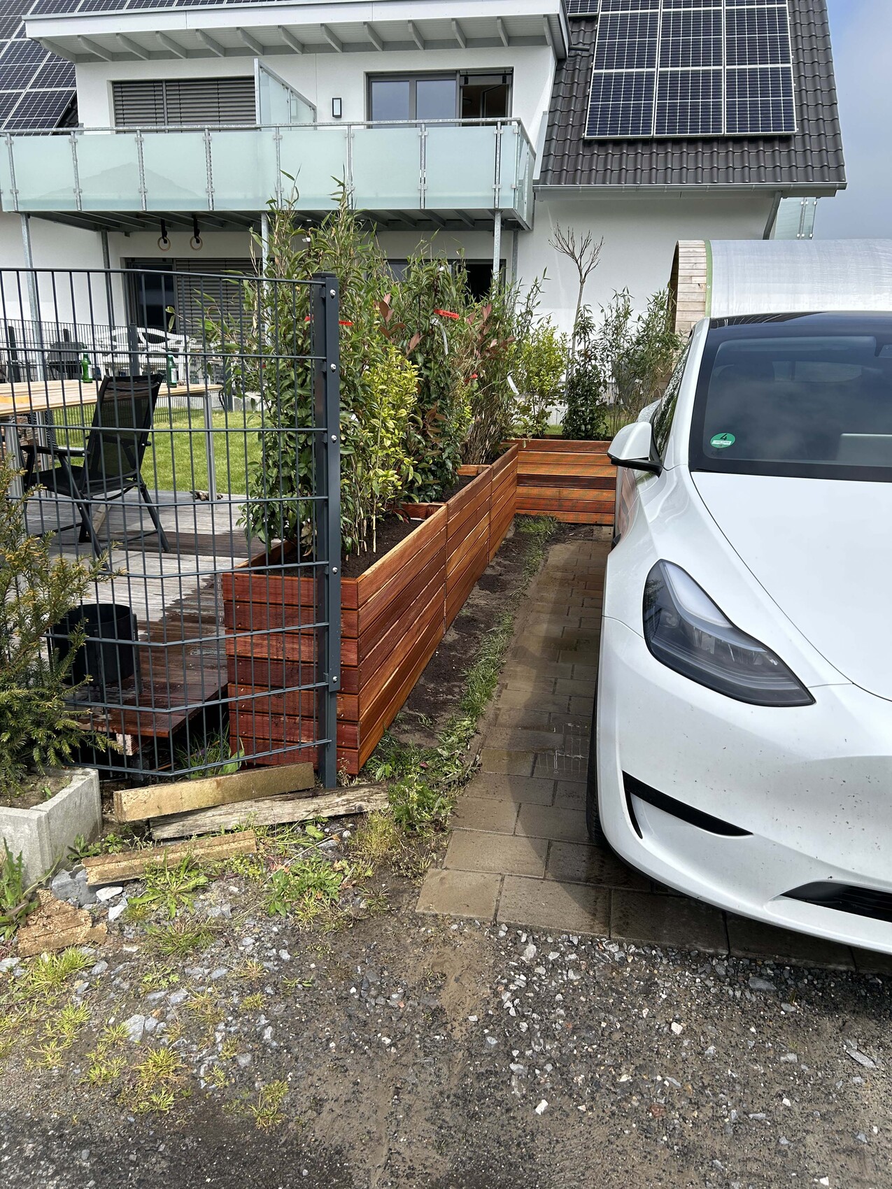 Modernes Haus mit Solaranlage, rechteckigen Holzbeeten, Garten und einem weißen Auto davor.