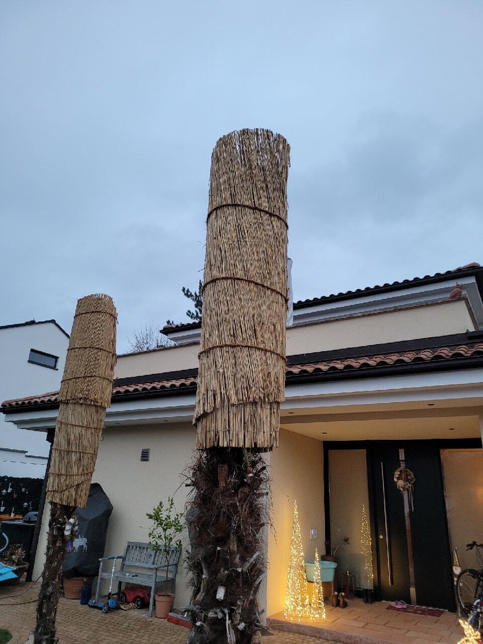 Zwei hohe Stroh-Säulen im Vorgarten eines modernen Hauses, Eingang mit Lichterkette.