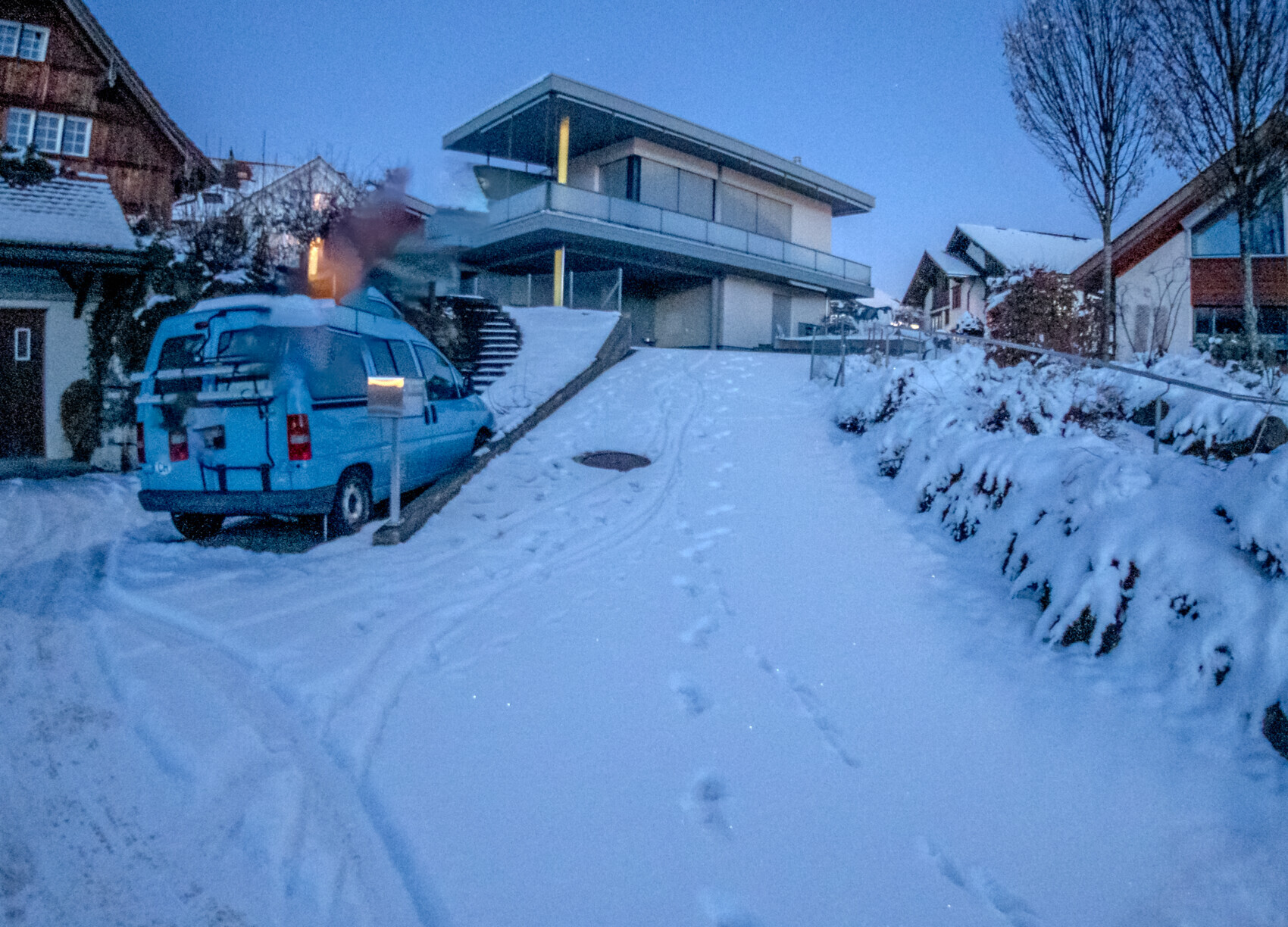 Blauer Lieferwagen steht auf einer schneebedeckten Straße vor modernen Häusern; Fußspuren im Schnee.