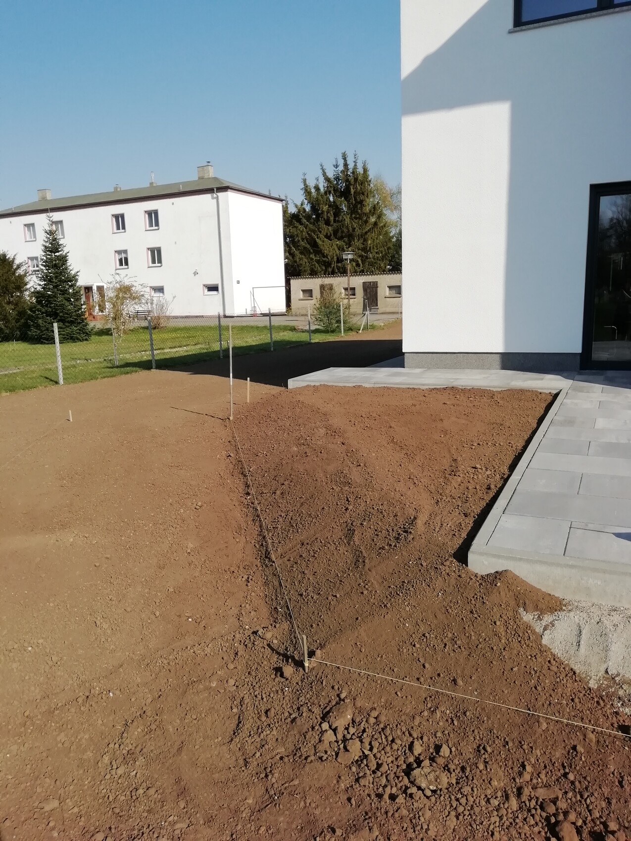 Baustelle neben modernem weißen Gebäude; Erdreich, Schnüre und Pfosten markieren Grundriss.