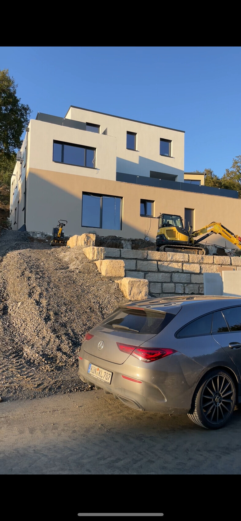 Modernes zweistöckiges Wohnhaus im Bau, Bagger, Kieshaufen und geparkter grauer Mercedes davor.