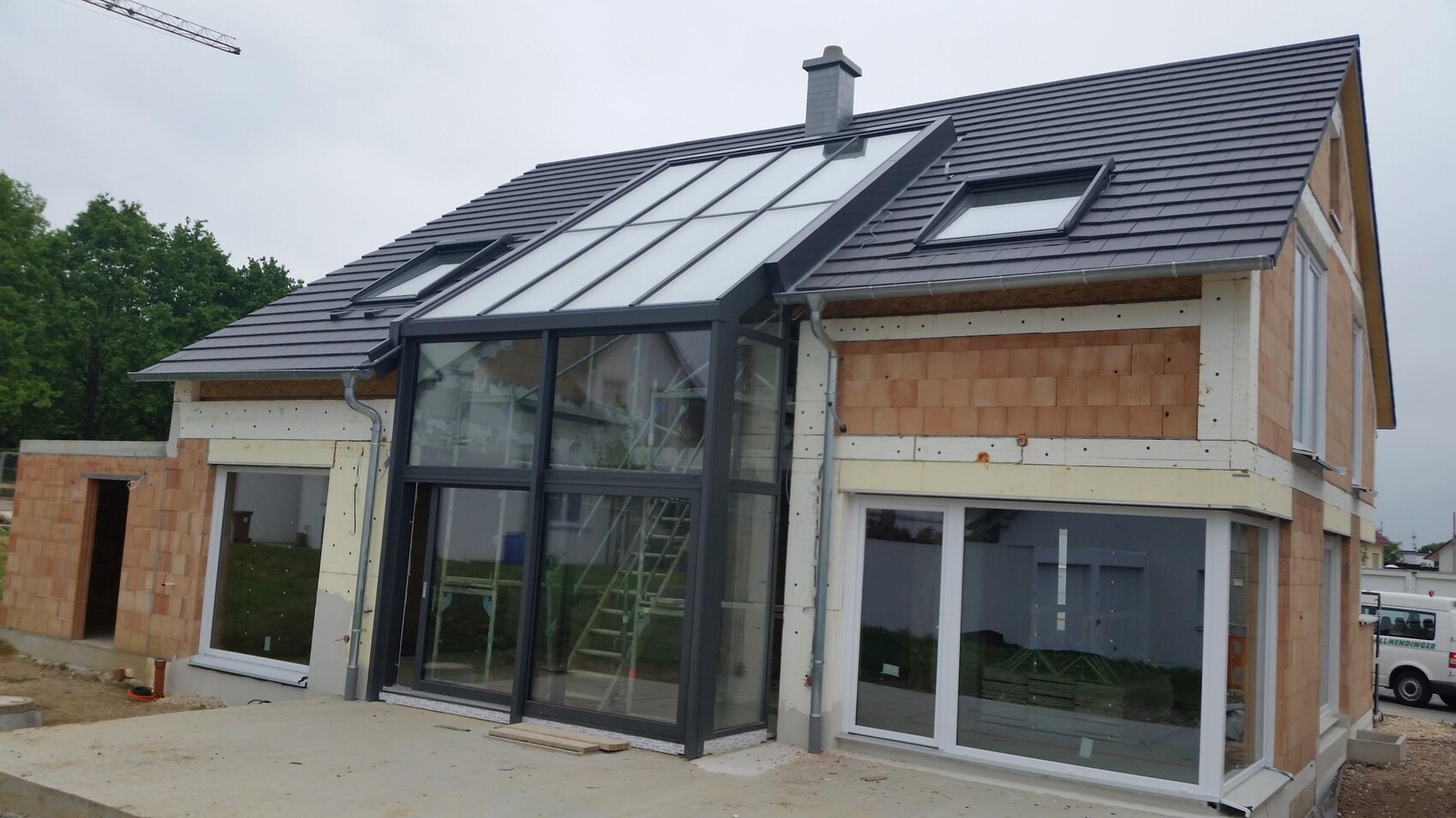 Neubau-Haus mit dunklem Ziegeldach und großer Glas-Wintergarten-Veranda; Baustelle sichtbar.