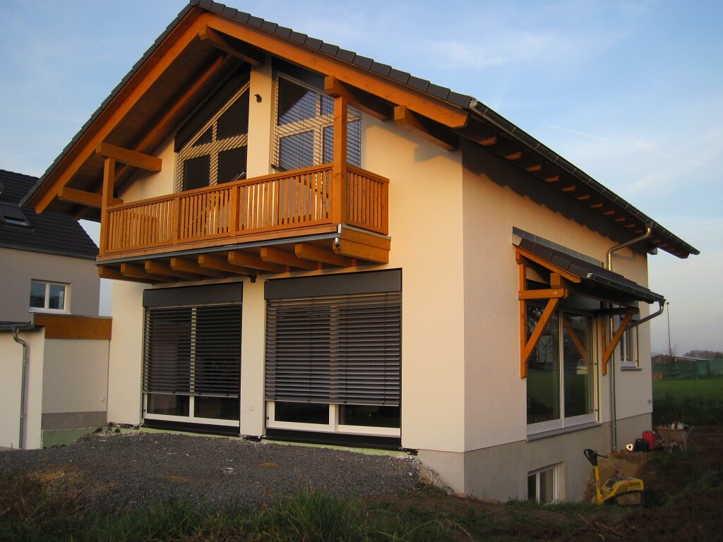 Zweistöckiges Haus mit Holzbalkon, großen Fenstern und Rollläden, Vorgarten.