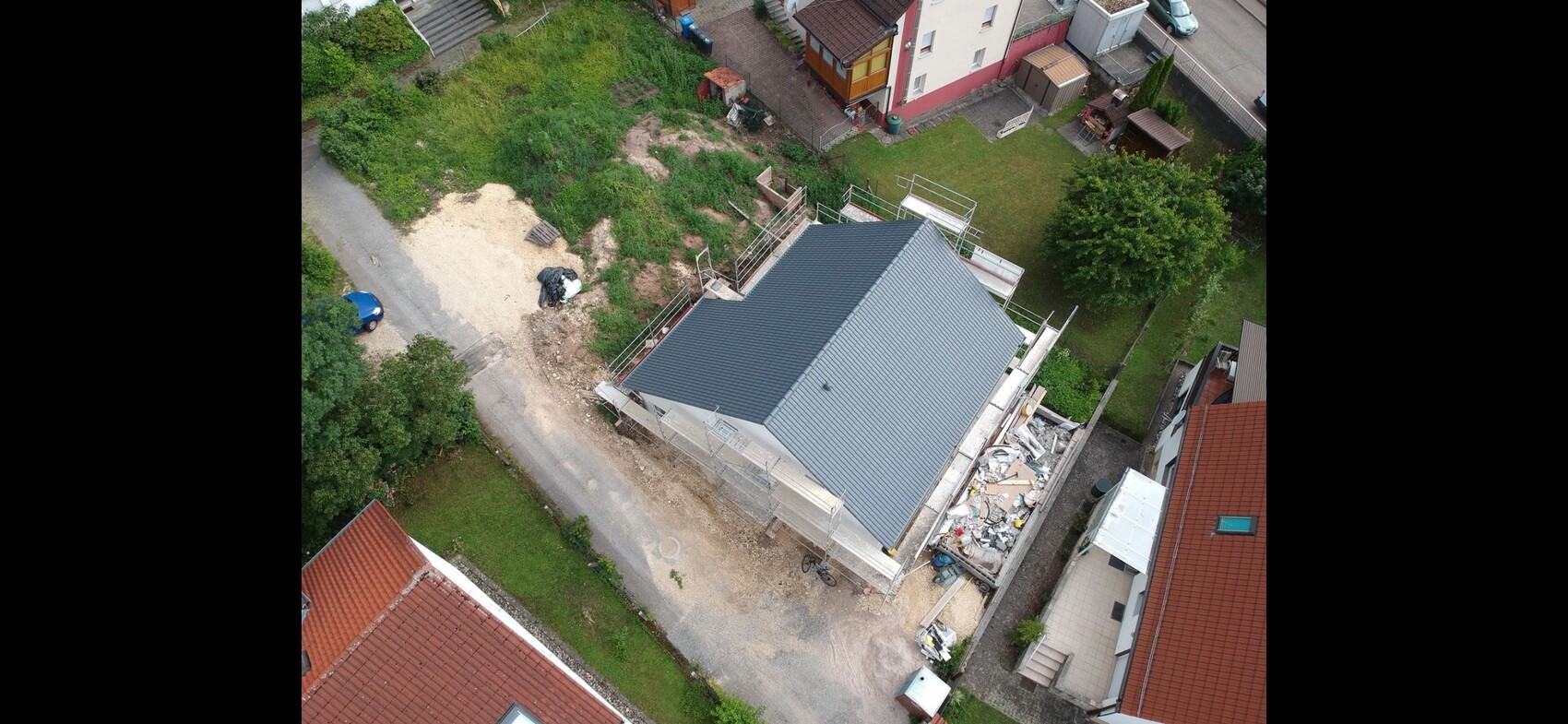 Vogelperspektive: Neubauhaus mit blauem Dach, Gerüst und Baustellenabfällen neben Garten.