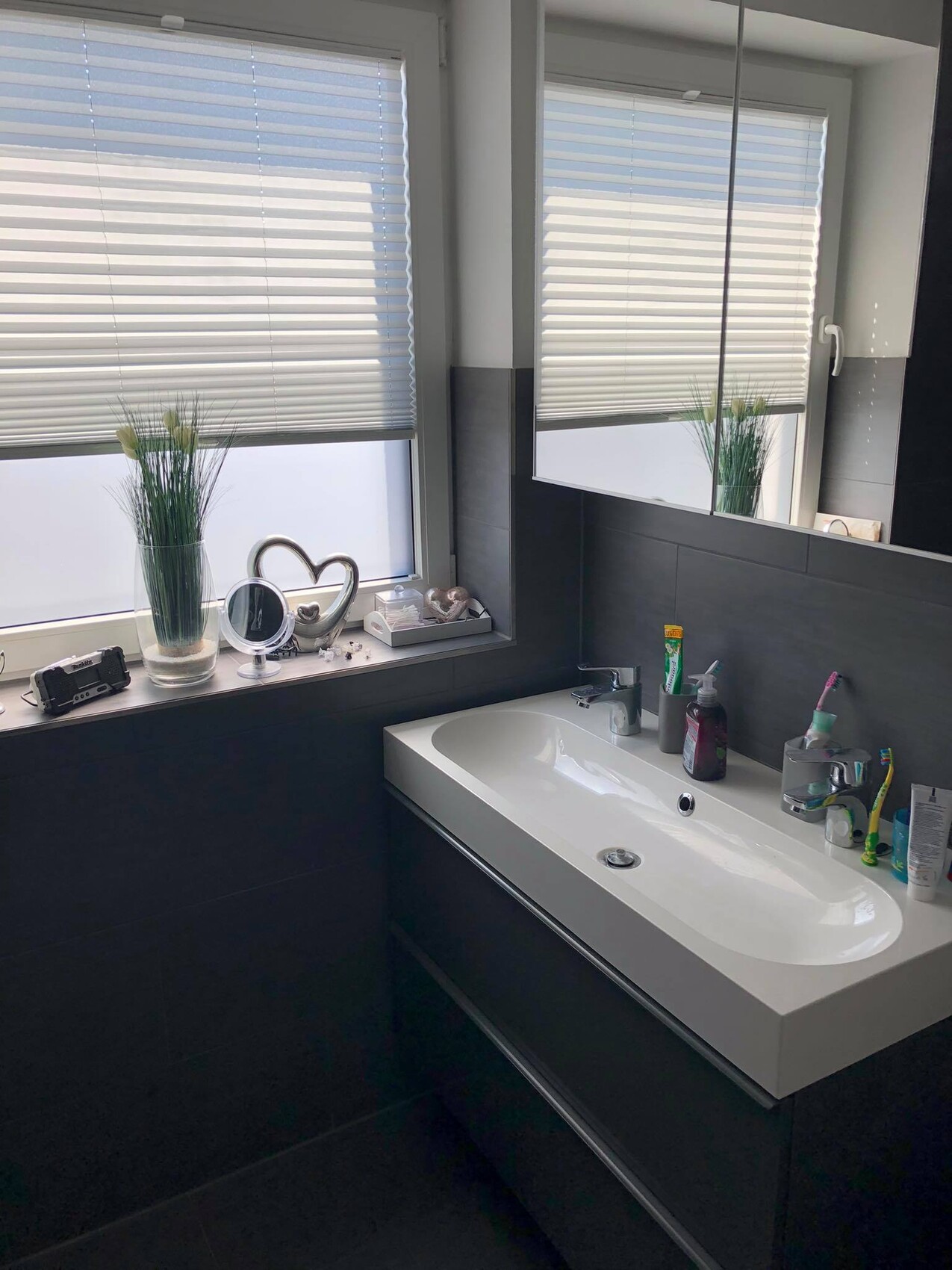 Badezimmer mit weißem Waschtisch, Spiegel, Fenster mit Jalousien und Deko auf der Fensterbank.