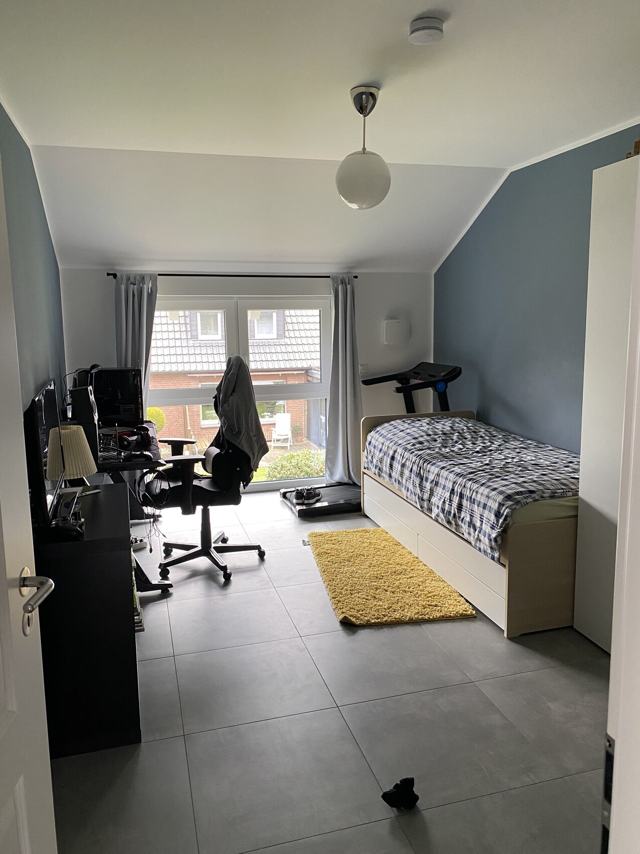 Schlafzimmer mit Bett, Schreibtischstuhl, Monitor, Laufband, Fenster mit Vorhängen.