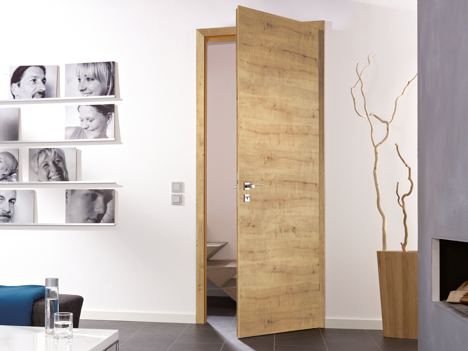 Helles Wohnzimmer mit offener Holztür, Wandgalerie schwarz-weiß Porträtfotos, Vase und Ast-Skulptur.