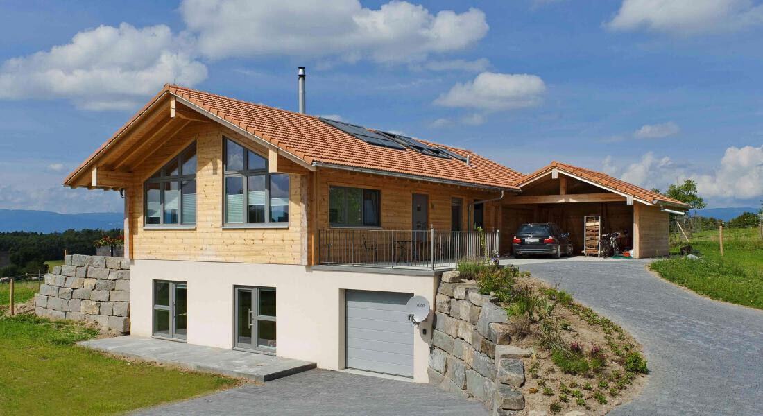 Modernes Holzhaus mit Carport, Garage, PV-Dach und Steinmauern auf Hanglage.
