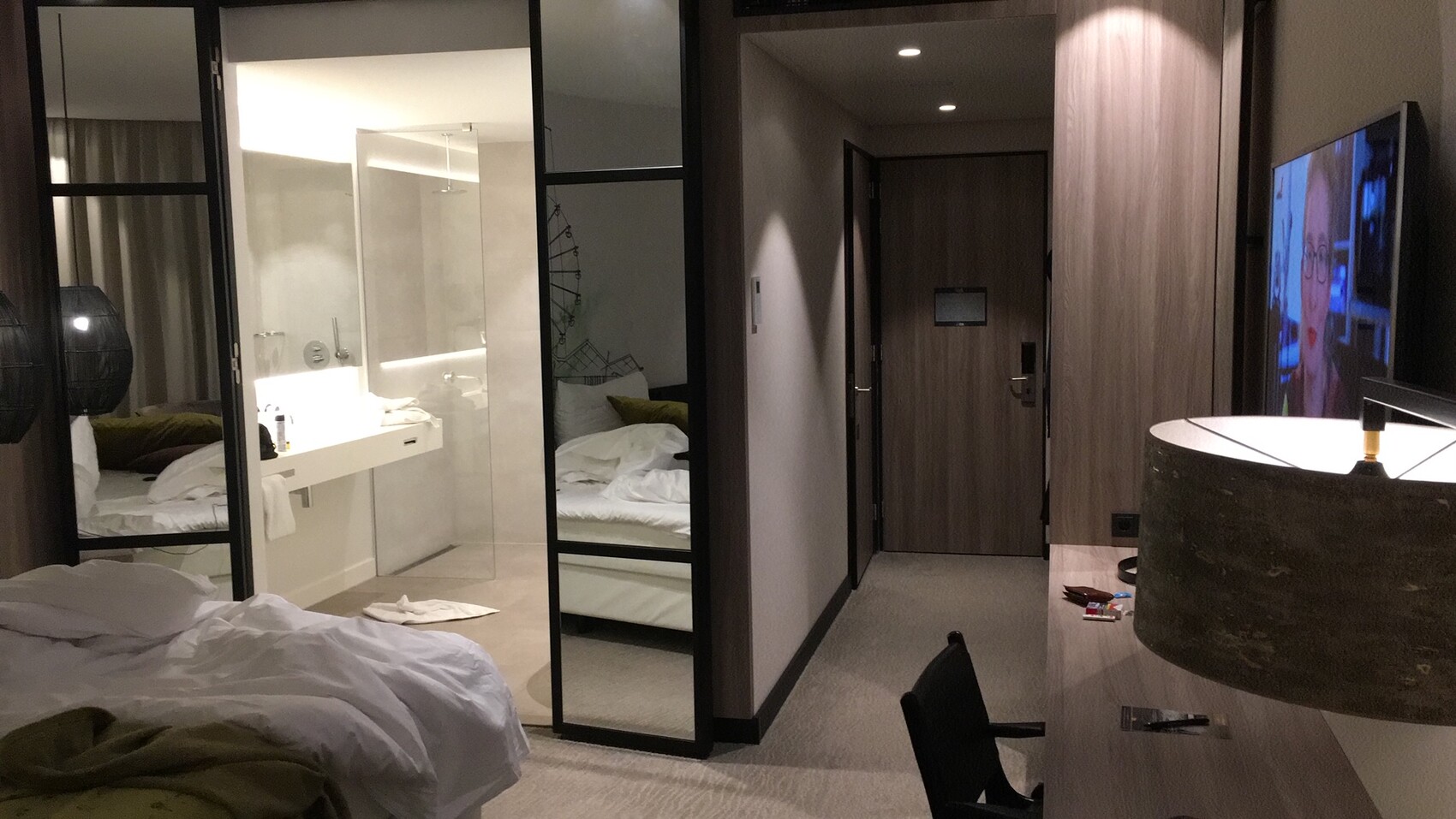Hotelzimmer mit Glaswand zum Bad, ungemachtes Bett, TV und Arbeitsbereich.
