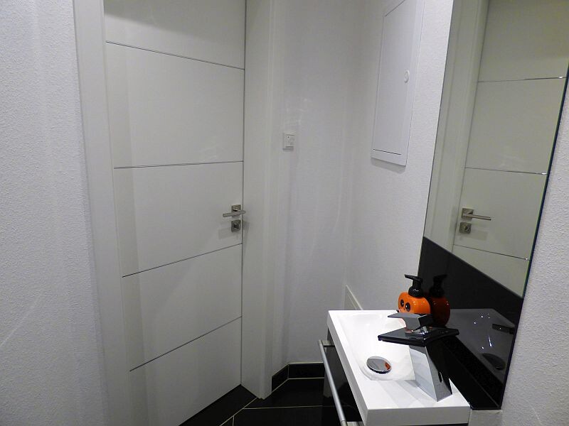 Weißes Badezimmer mit Waschbecken, Spiegel, orange Seifenspender und weißer Tür.