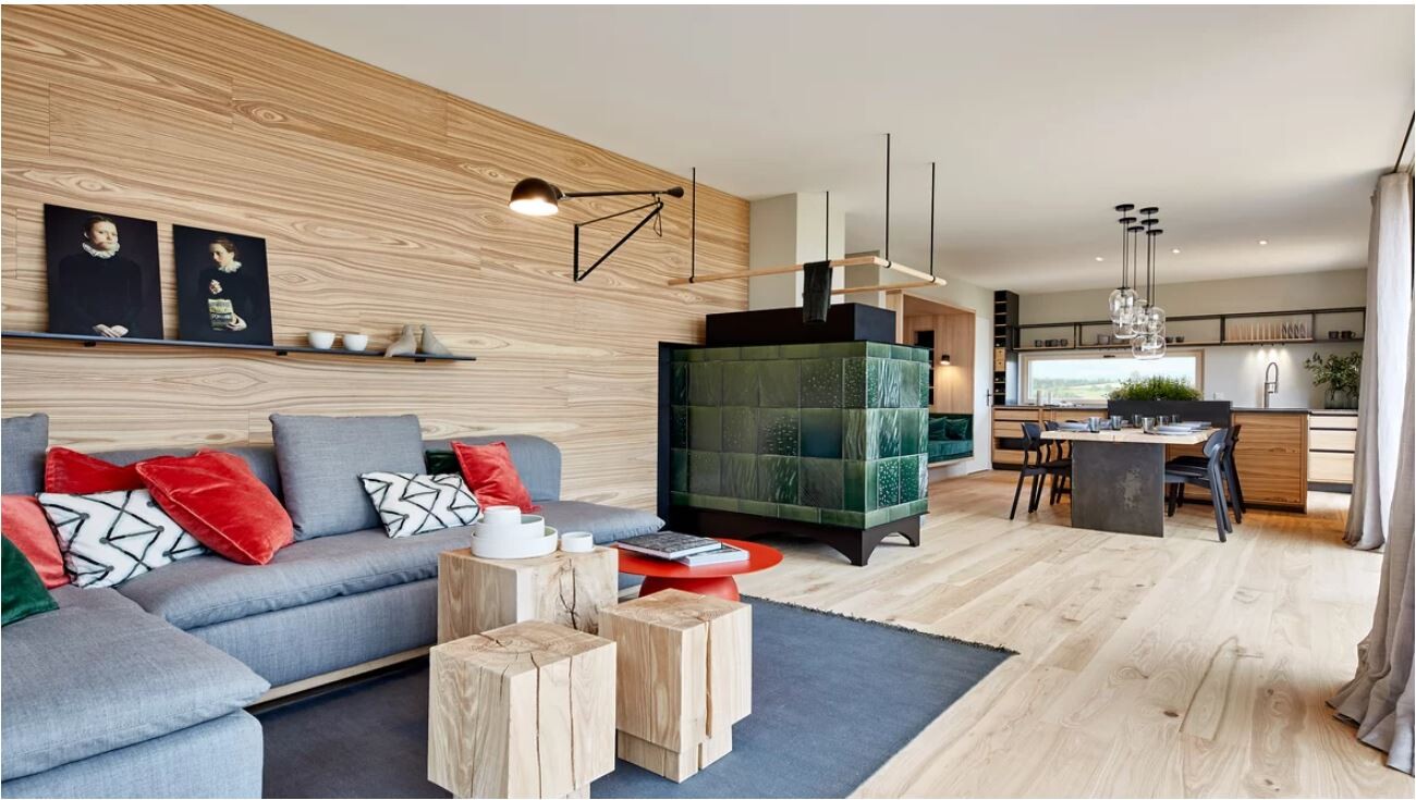 Wohnzimmer mit grauem Sofa, Holzpaneelwand, grünem Kachelofen, Esstisch und Küche im Hintergrund.
