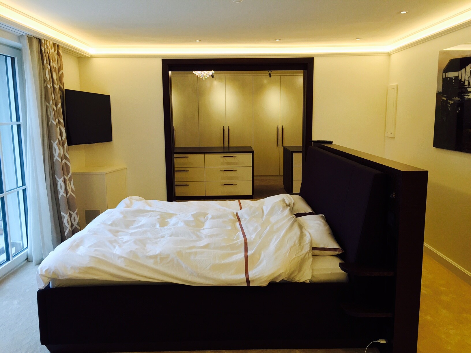 Schlafzimmer: dunkles Bettgestell, weiße Bettwäsche, TV links, begehbarer Kleiderschrank
