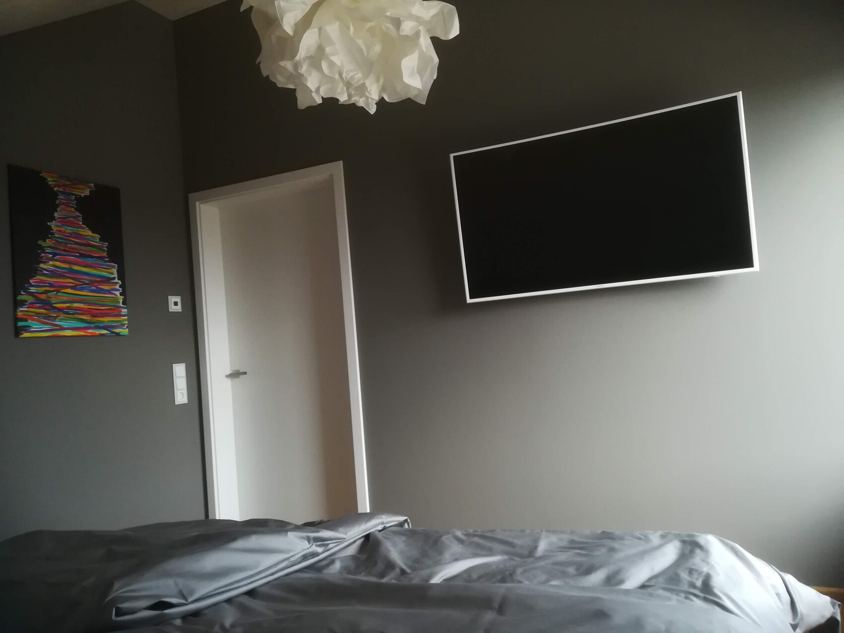 Graues Schlafzimmer, schräg montierter Flachbild-TV an der Wand, offene Tür, buntes Kunstwerk links.