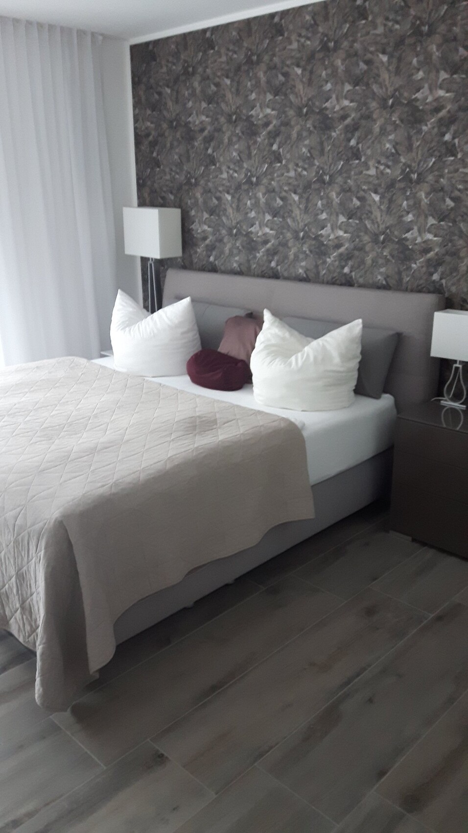 Bett mit beige-Quilt, weißen Kissen, dunkle gemusterte Wand und Nachttischlampen.