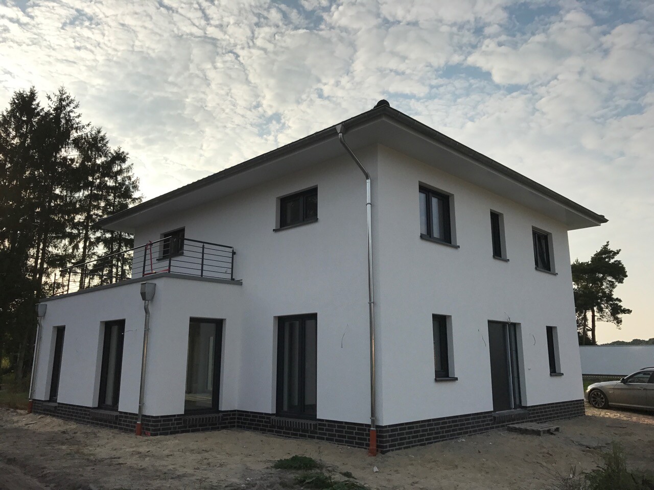 Ein weißes, zweigeschossiges Haus mit Balkon, dunklem Sockel, Baugrund und einem Auto daneben.