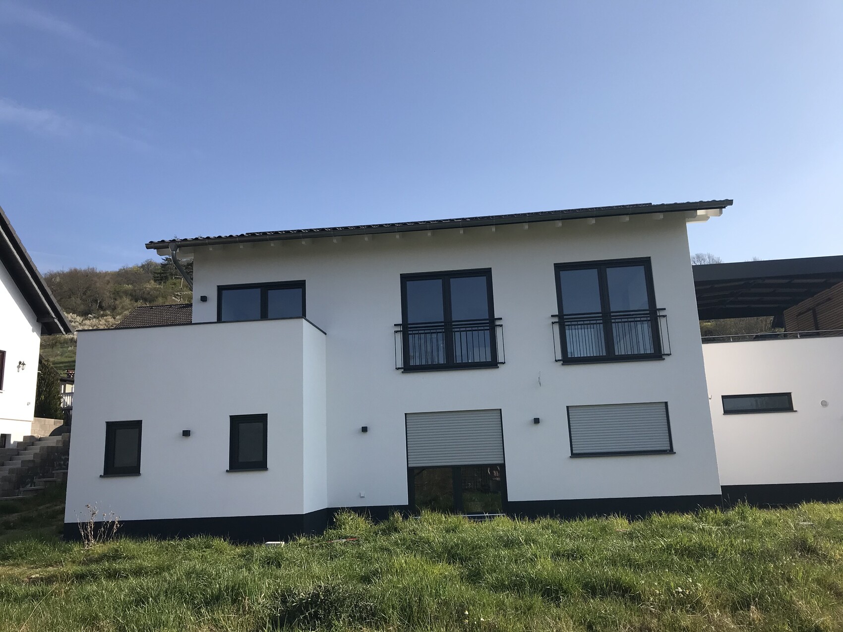Modernes weißes Einfamilienhaus mit zwei Balkonen, großen Fenstern, dunklem Sockel und Grasfläche.
