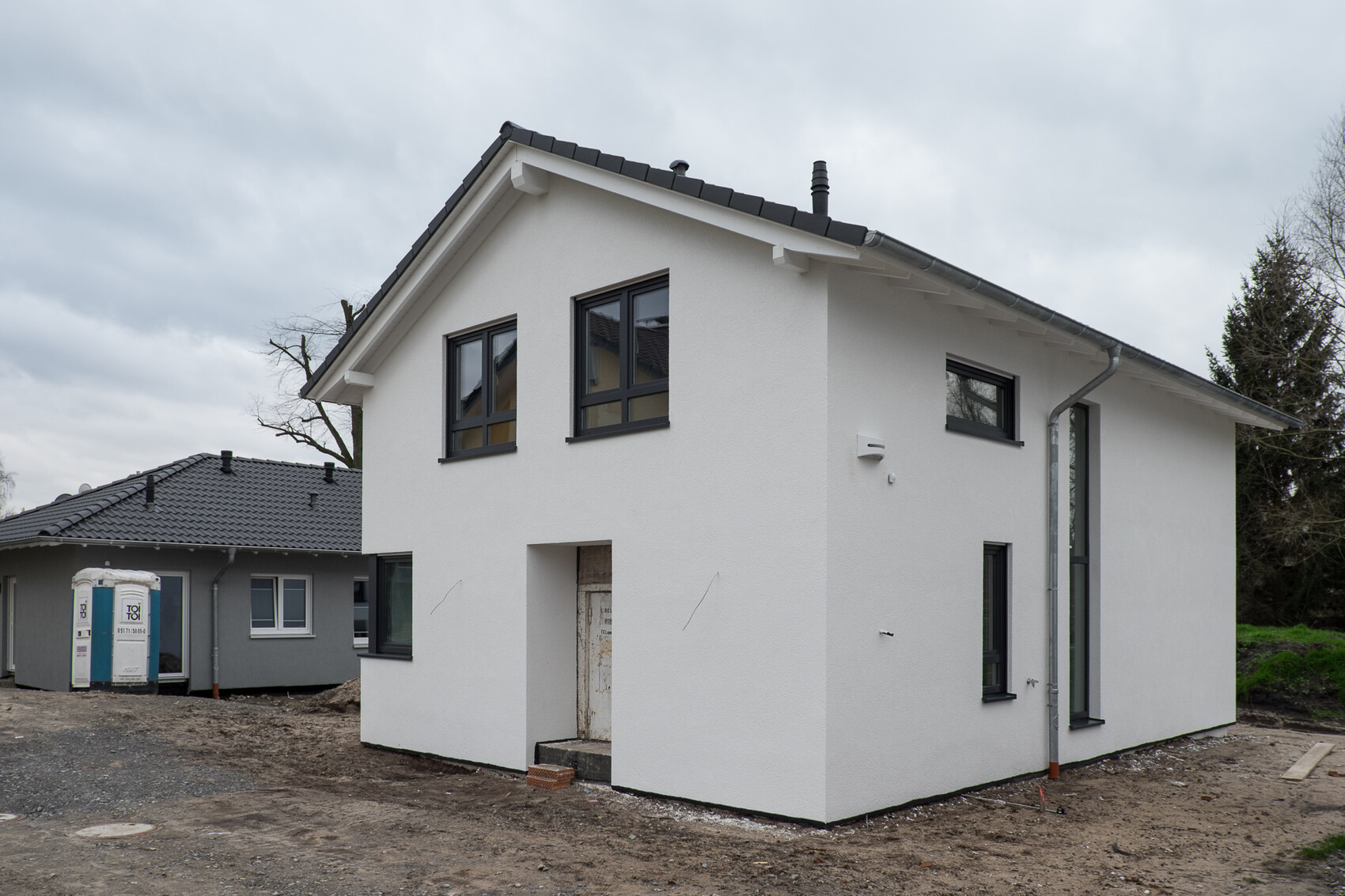 Weiße moderne Hausfront, zweistöckig, graue Fensterrahmen, Baustelle mit Porta-Potti im Hintergrund.
