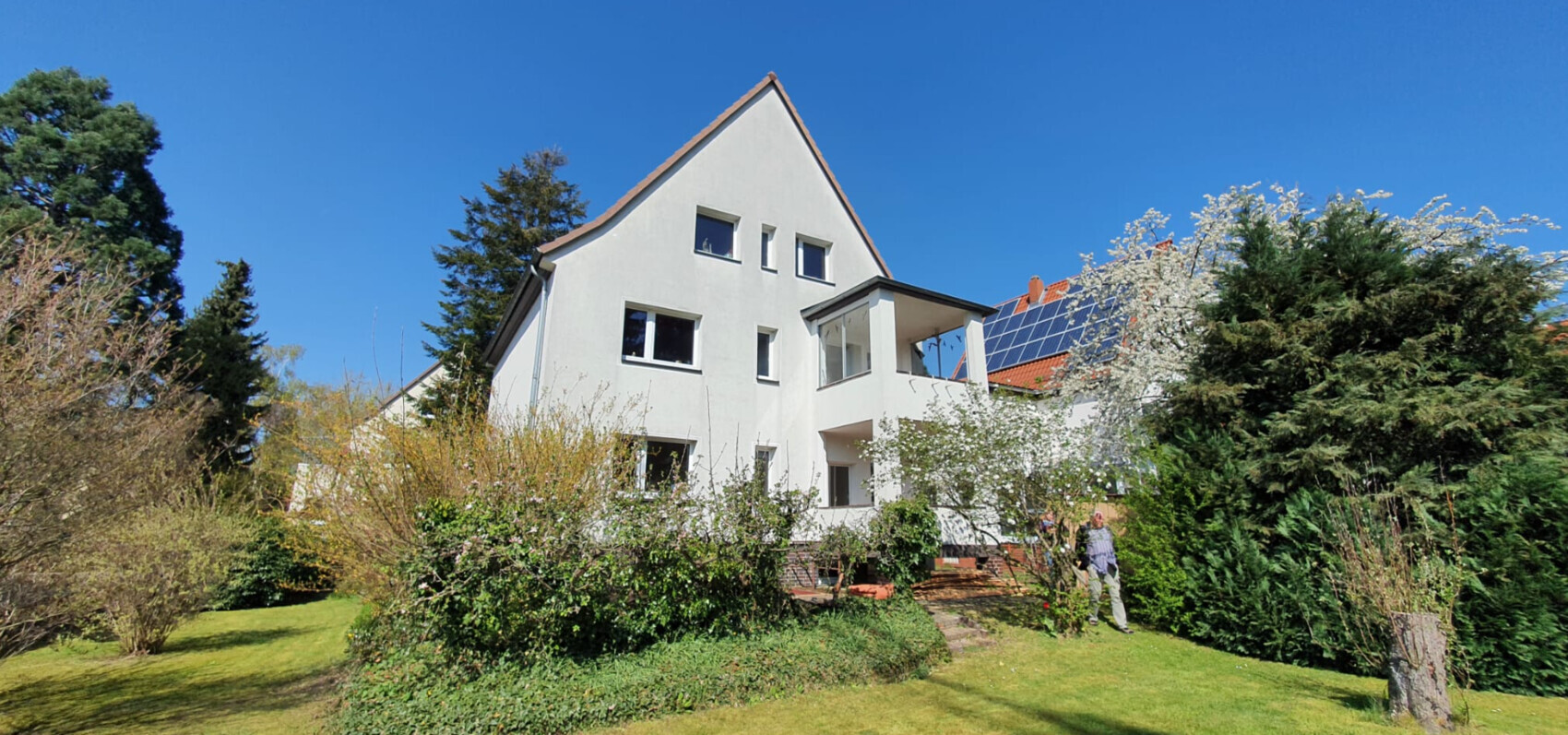 Weißes mehrstöckiges Haus mit Garten, Bäumen und Solarpanelen auf dem Dach
