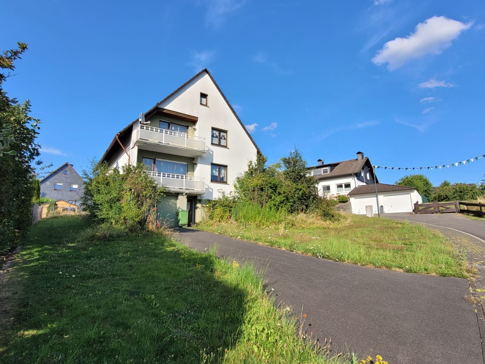 Weißes Mehrfamilienhaus mit steilem Dach und Balkonen, Garten, Straße und blauem Himmel.