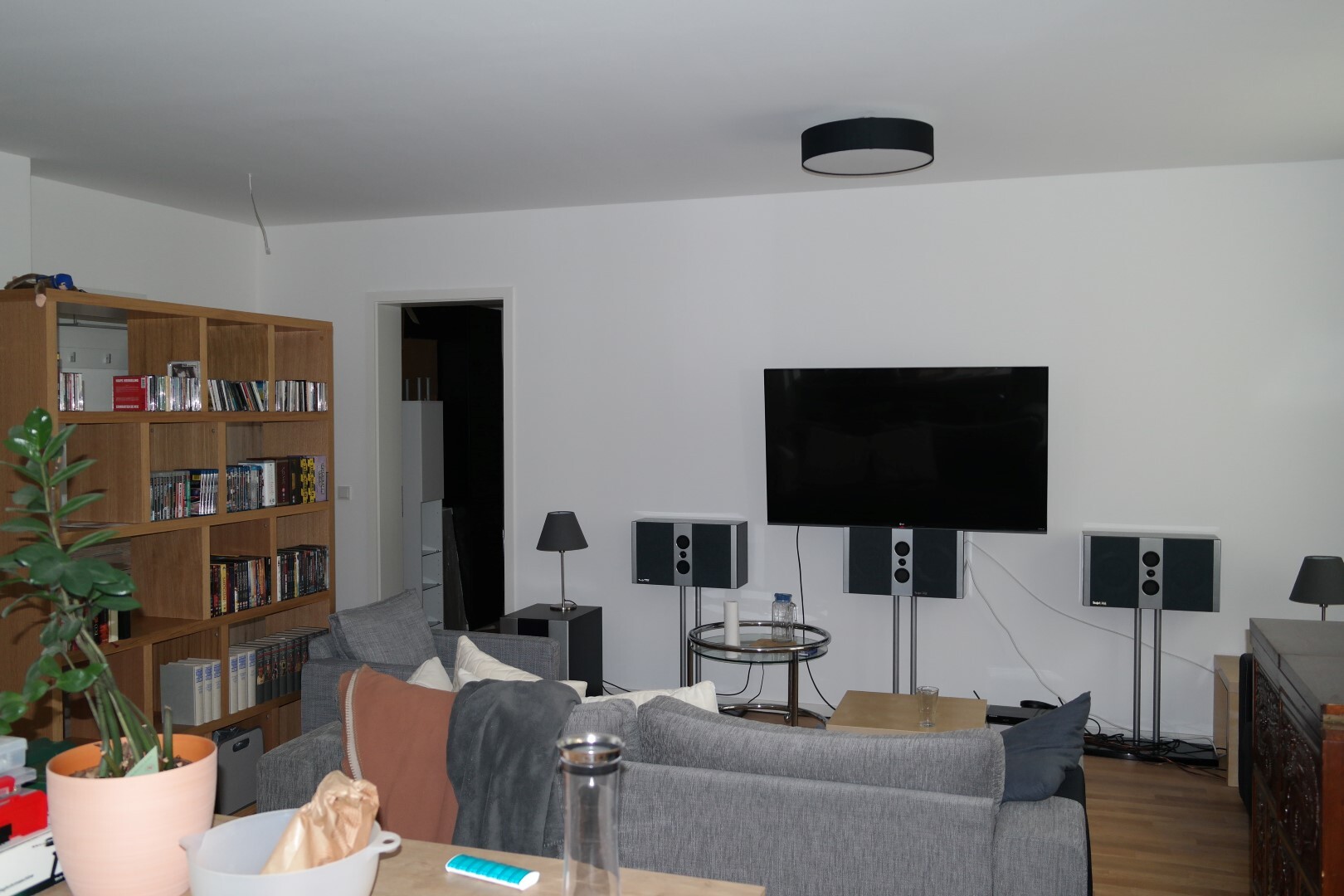 Wohnzimmer mit Großbild-TV an weißer Wand, Surround-Soundsystem, Sofa und Bücherregal.