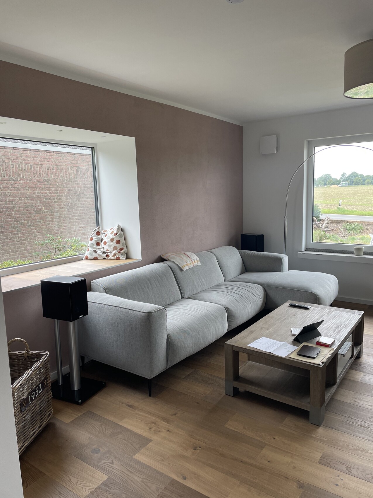 Helles Wohnzimmer mit grauer Eckcouch, Holztisch, großen Fenstern, brauner Akzentwand und Arc-Lampe.