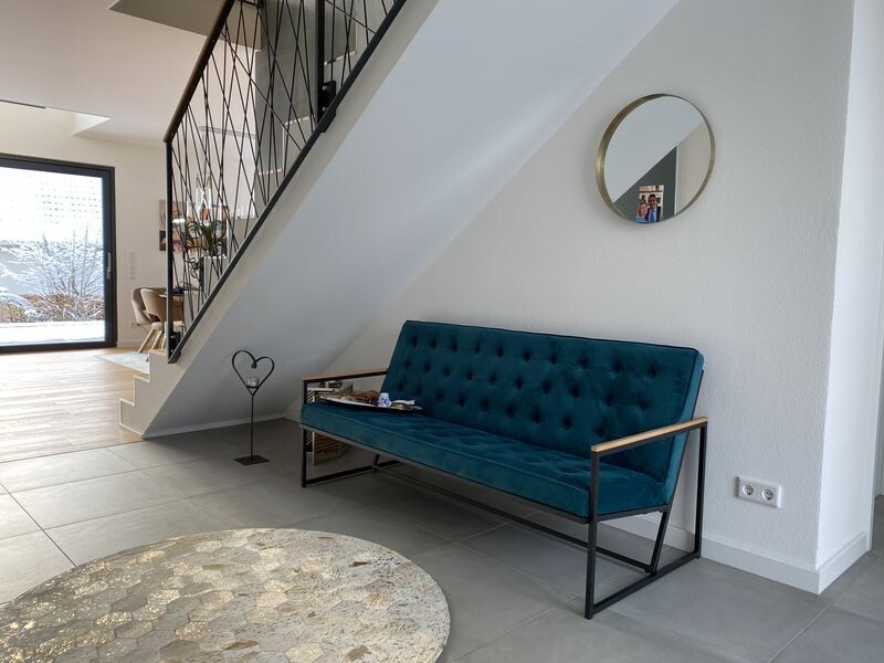 Modernes Wohnzimmer mit tealfarbener Couch unter der Treppe und rundem Spiegel an weißer Wand.