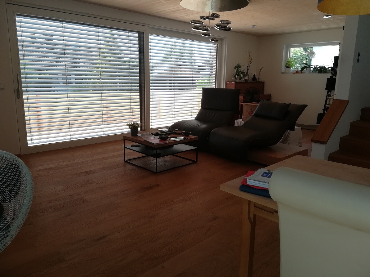 Wohnzimmer mit Holzboden, große Fensterfront mit Jalousien, zwei schwarze Relaxsessel.