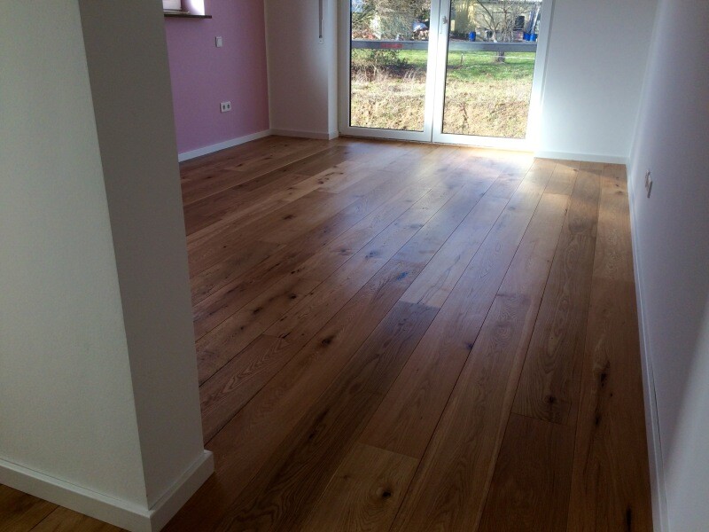 Leerer Wohnraum mit Holzboden, pinker Wand links, weißen Wänden und großen Glasfenstern am Ende.