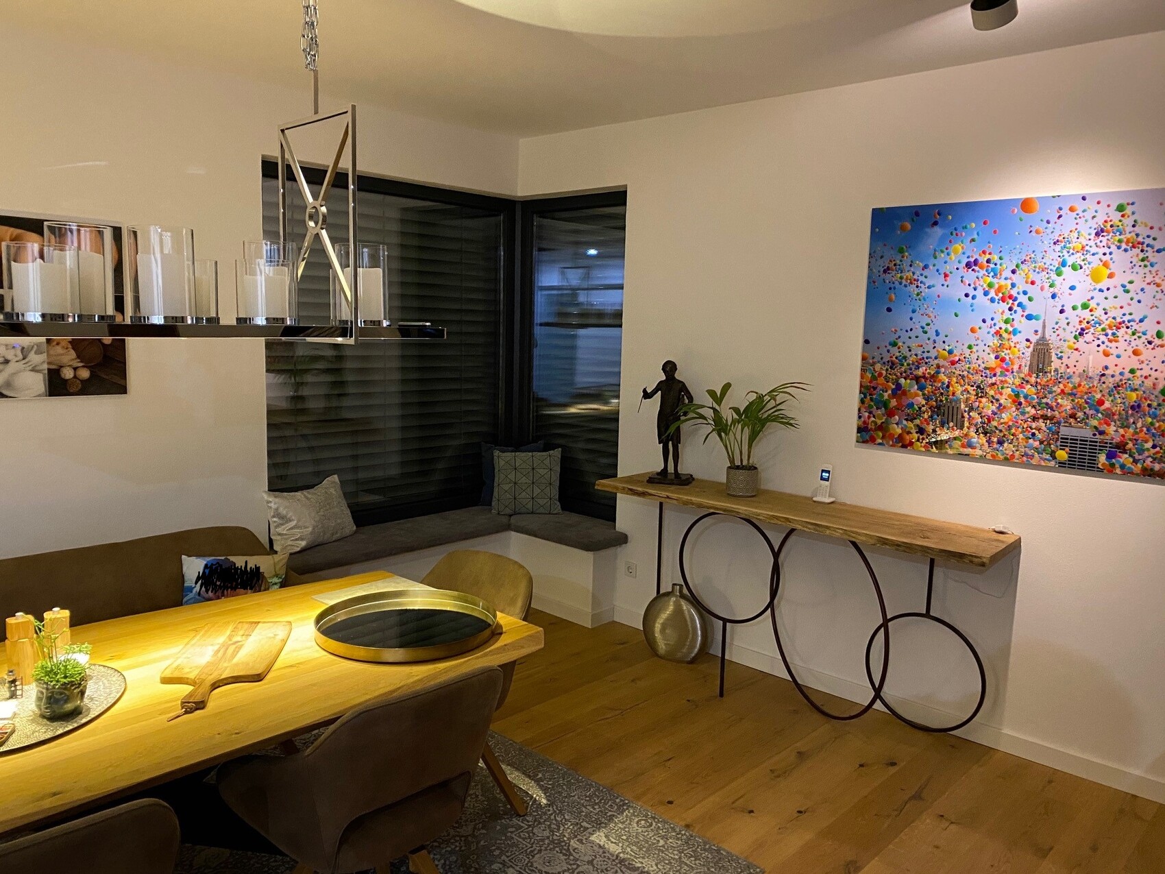 Helles Esszimmer mit Holztisch, Eckbank, moderner Leuchter, Pflanze und bunter Stadt-Gemälde.