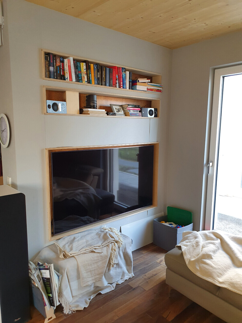 Wohnzimmer mit Wand-TV in Holzrahmen, darüber Bücherregale; Sofa mit Decke.