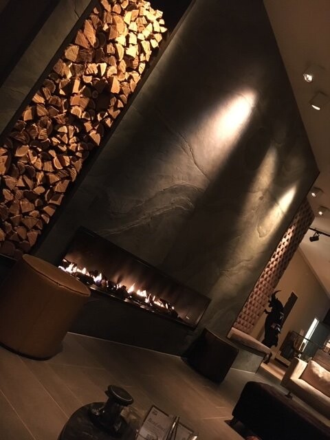 Moderner Wohnraum mit langem Kamin, Flammen, links gestapeltem Holz und dunkler Wand.