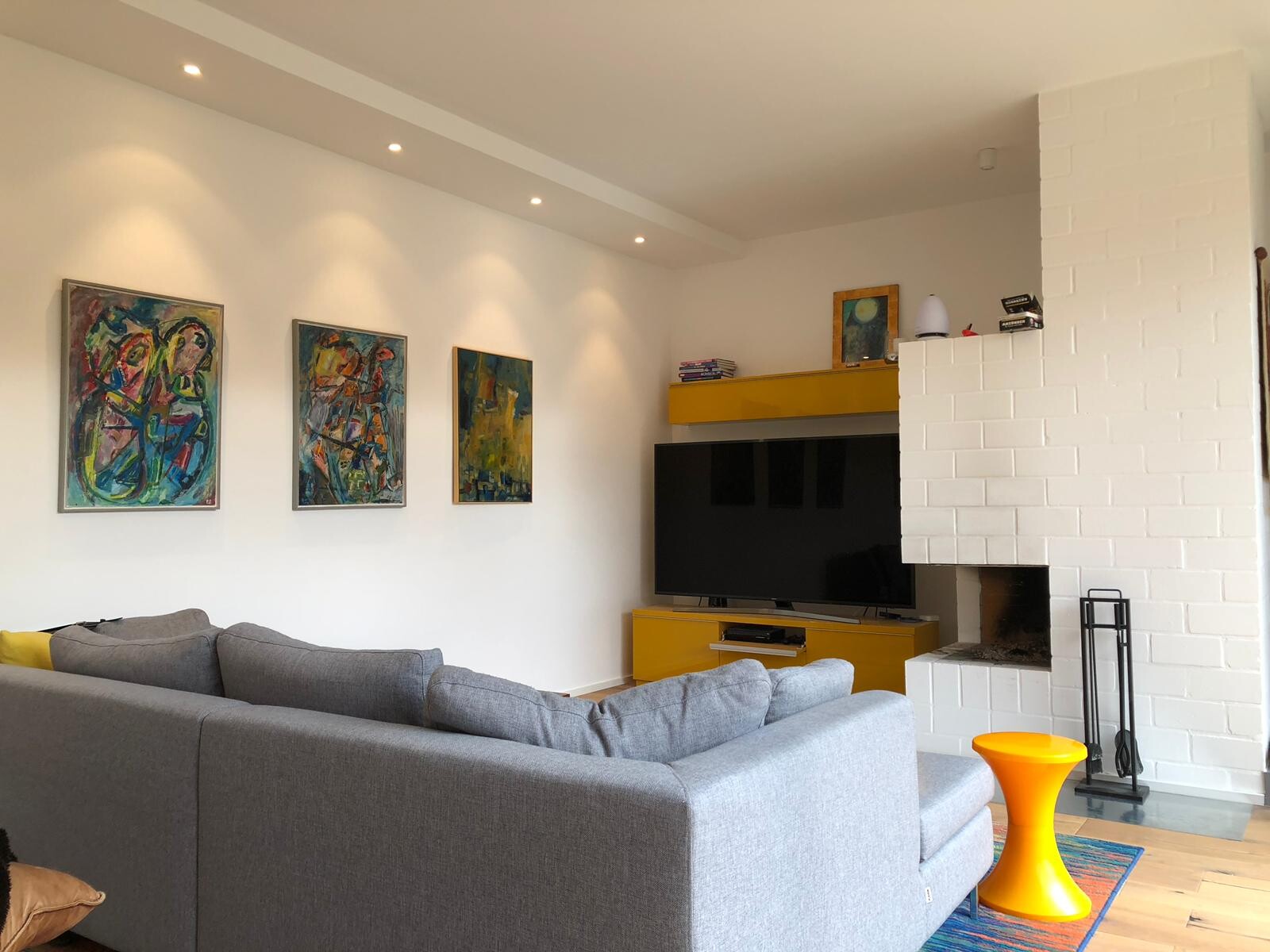Wohnzimmer mit grauer Eckcouch, bunten abstrakten Bildern und gelber TV-Konsole.