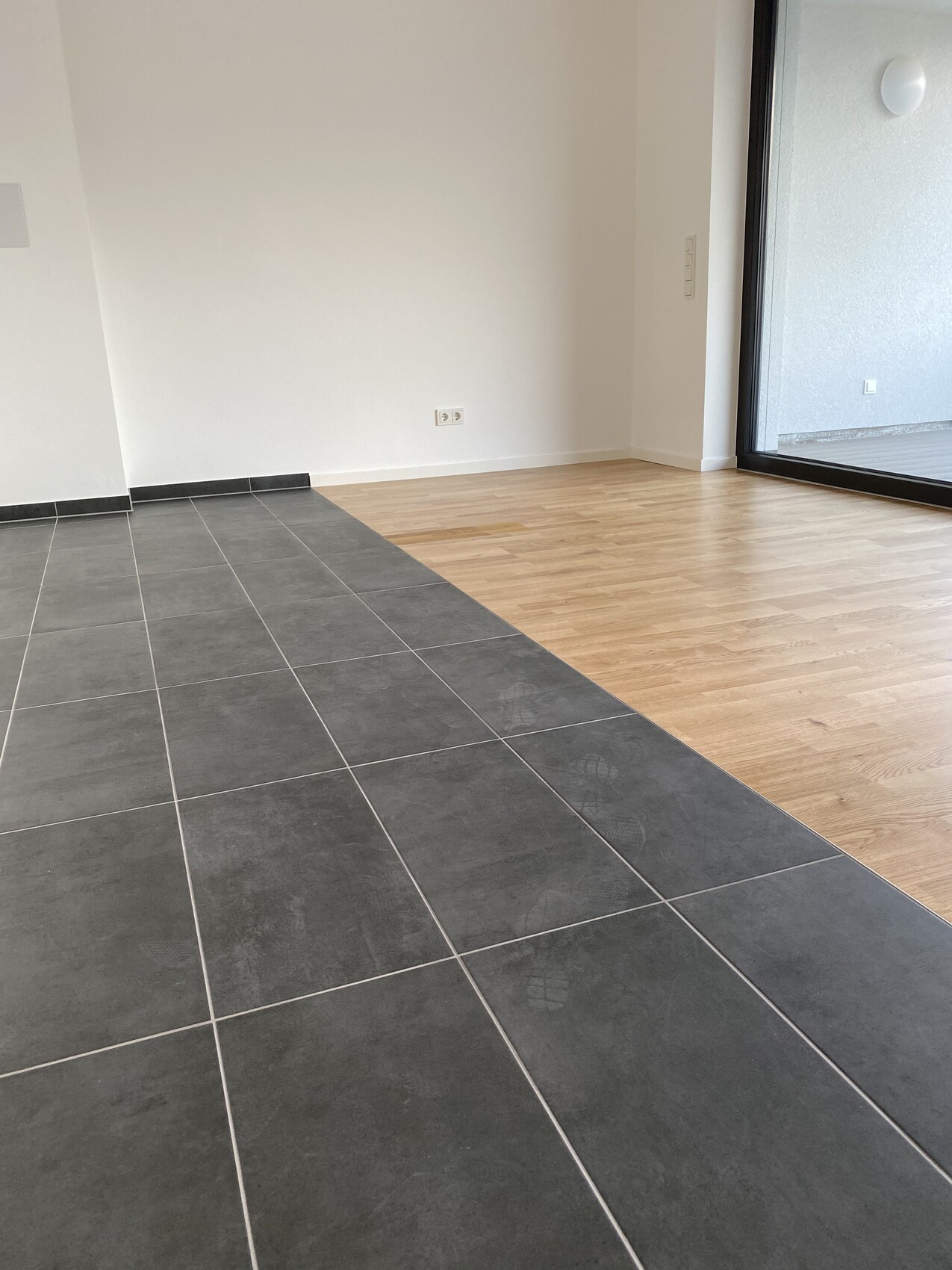 Moderne Innenansicht: dunkle Fliesen links, Holzboden rechts, Glastür nach außen.