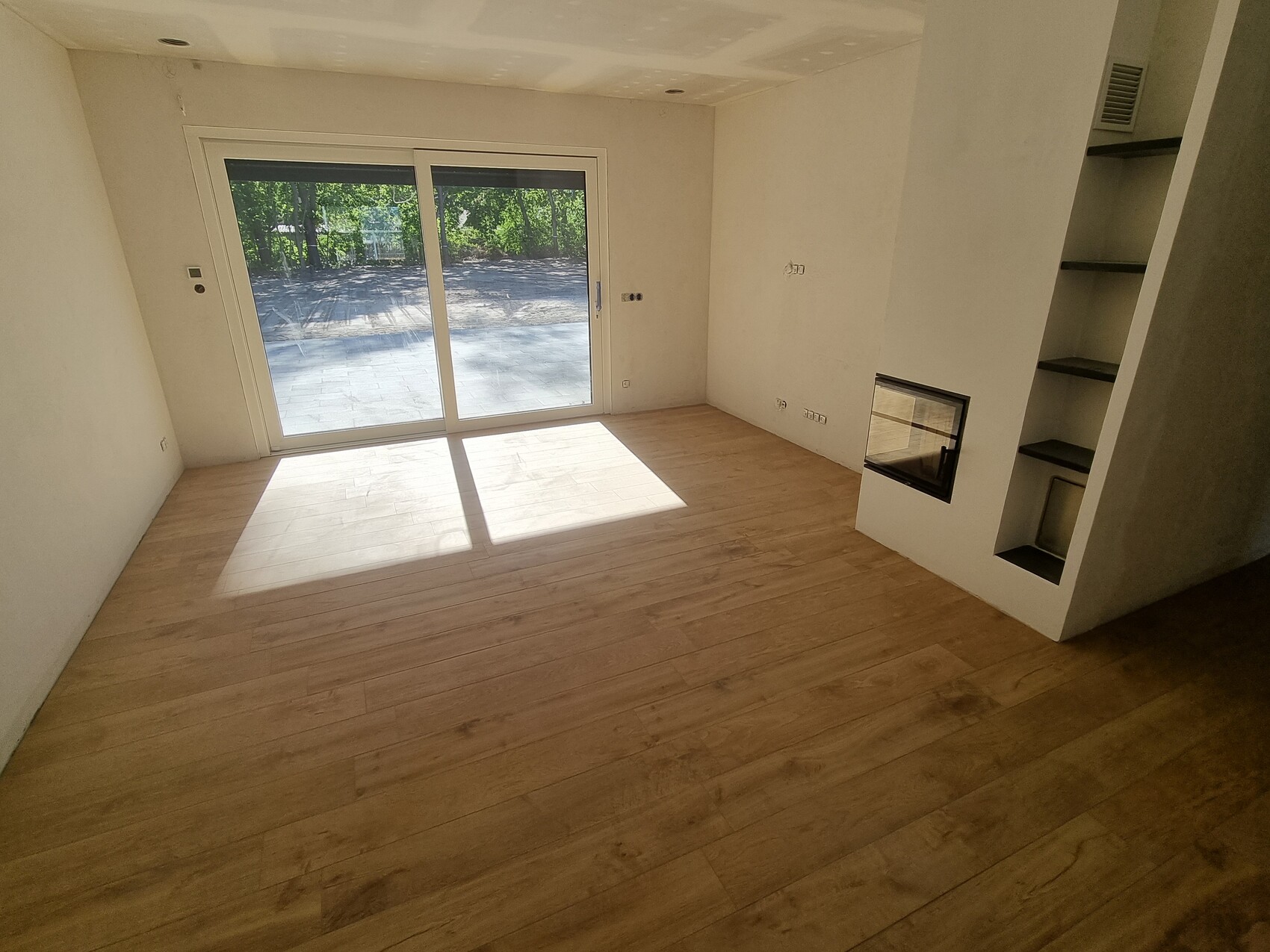 Leerer Wohnraum mit Holzfußboden, Glas-Schiebetür zum Garten, Kamin-Nische mit Regalen.