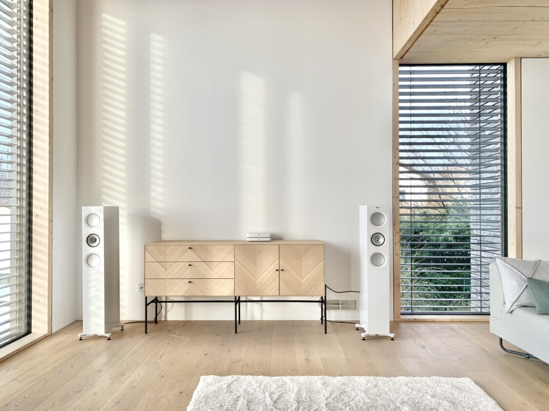 Modernes Wohnzimmer mit Holzsideboard, weißen Standlautsprechern und großen Fenstern.
