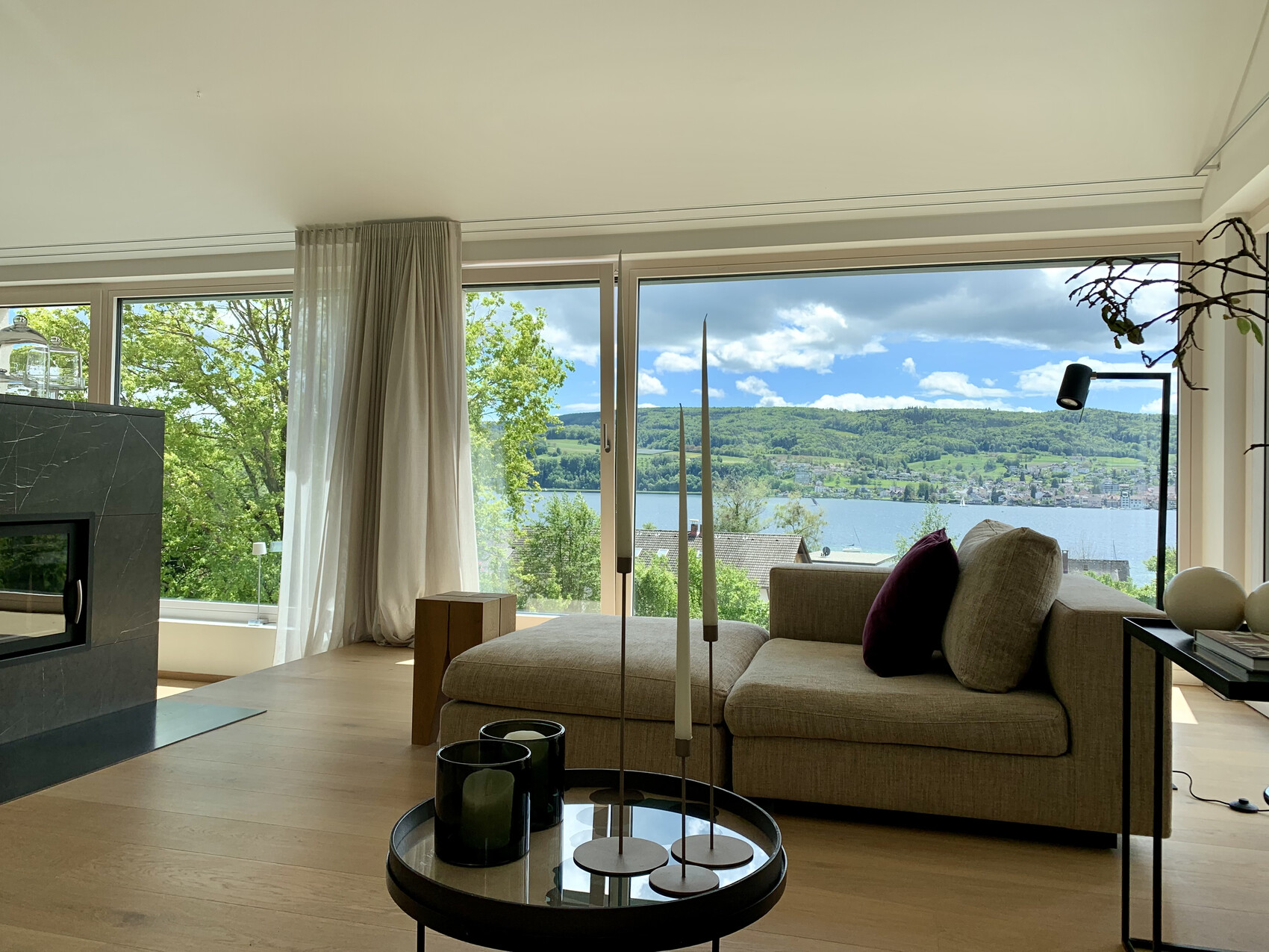 Geräumiges Wohnzimmer mit Großfenstern, Blick auf See und Berge, Kamin links, Sofa.