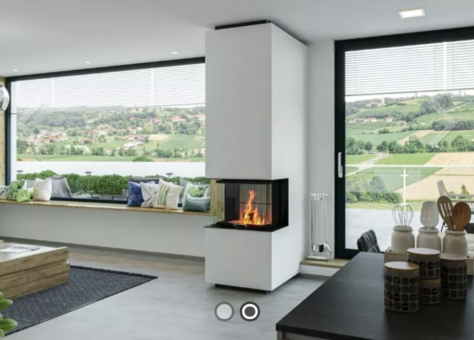 Offenes Wohnzimmer mit freistehendem Kamin, Fensterbank mit Kissen und Panoramablick auf grüne Umgebung.