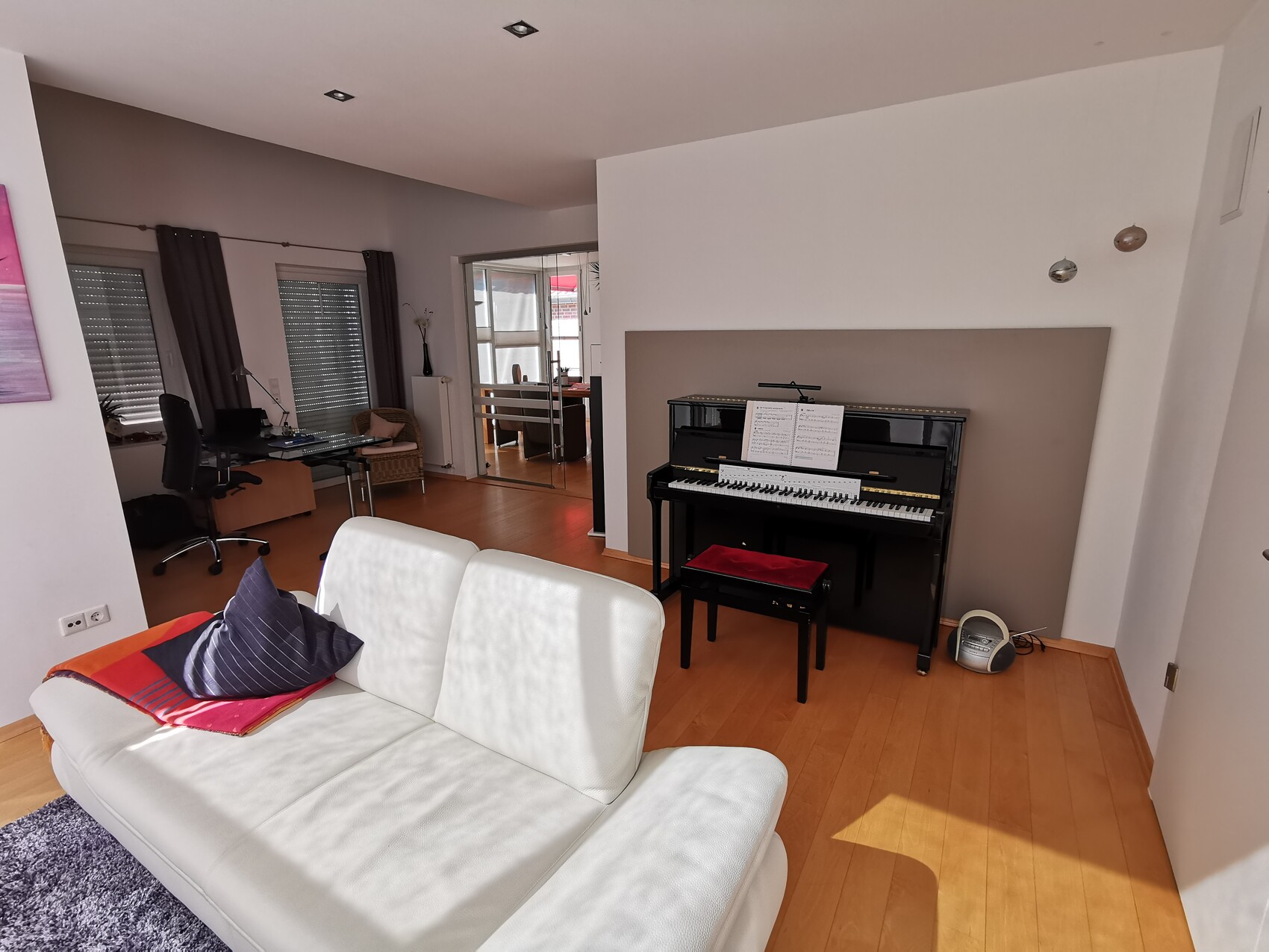 Wohnzimmer mit weißem Sofa, schwarzen Klavier, Notenblatt, Arbeitsbereich links, Holzboden.