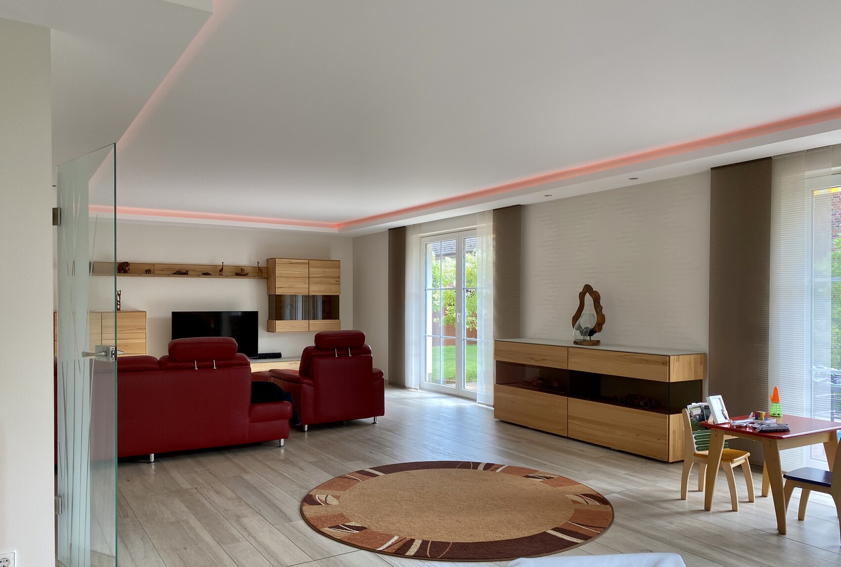 Offenes Wohnzimmer mit roten Ledersofas, Holzschränken, Glasdurchgang, runder Teppich, Spieltisch.