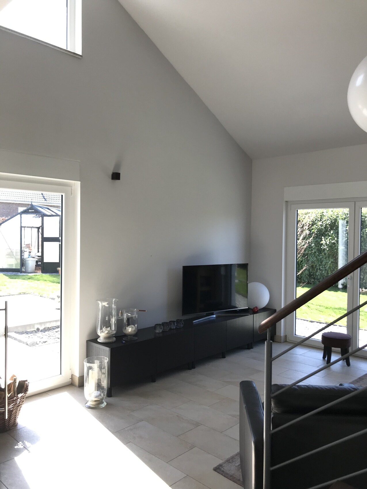 Modernes Wohnzimmer mit Fernseher, dunkelgraues Sideboard, Treppe und grossen Glasfenstern.