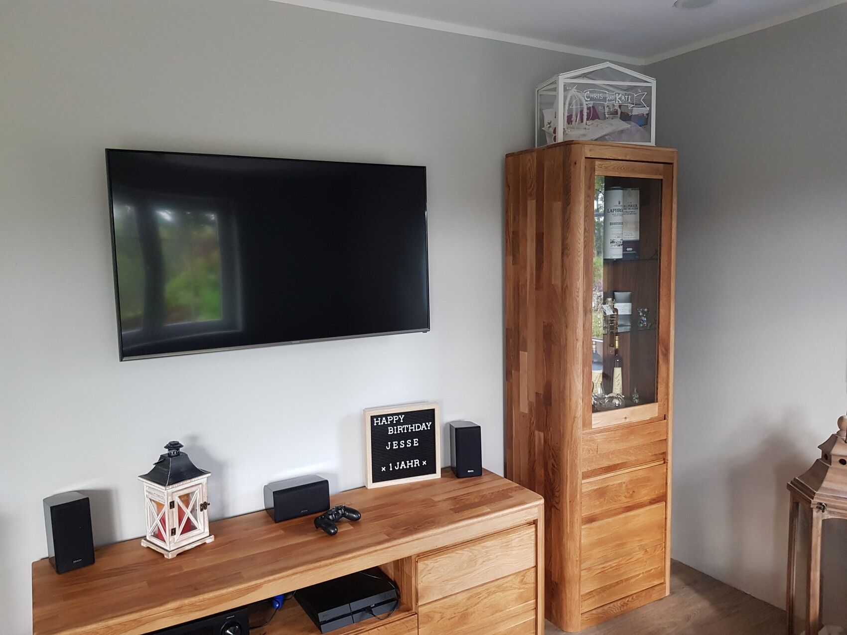 Wohnzimmer-Ecke mit Wandfernseher, Holzsideboard, Glasvitrine mit Flaschen, Geburtstagsanzeige.
