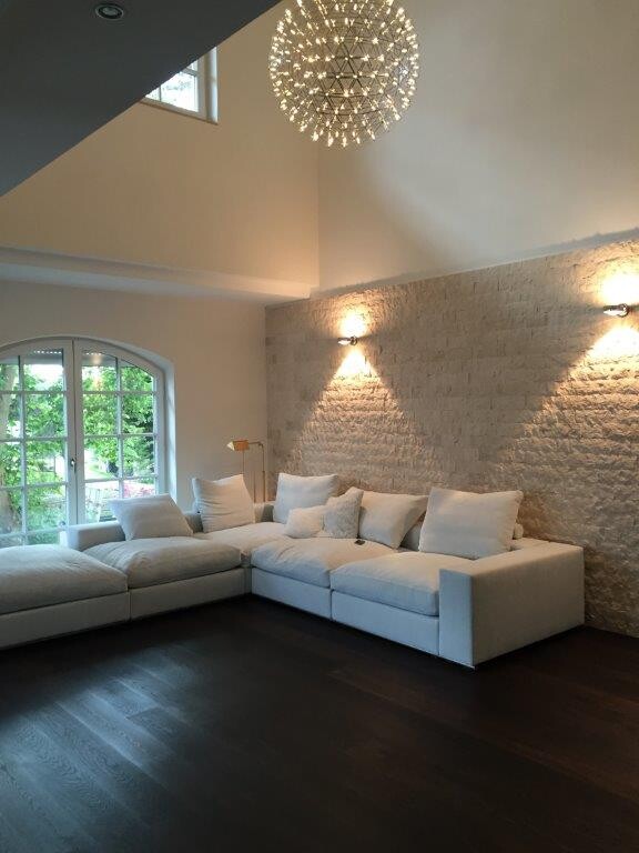 Modernes Wohnzimmer mit weißem Ecksofa vor Steinwand, dunklem Holzboden, Bogenfenster und Kugelleuchte.