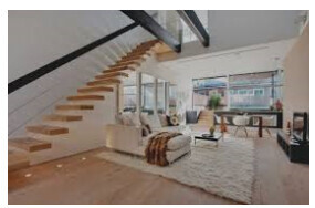 Modernes Wohnzimmer mit offener Treppe, großen Fenstern und gemütlicher Sitzecke auf hellem Holzboden.
