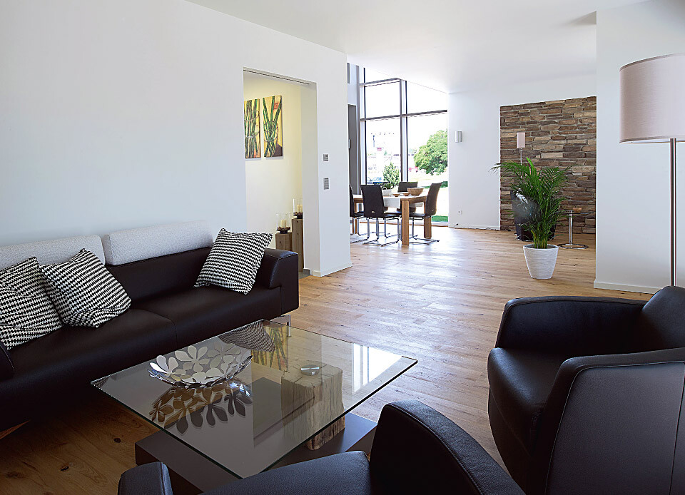 Offenes Wohnzimmer mit schwarzer Ledercouch, Glas-Tisch, Holzboden und Essbereich vor großen Fenstern.