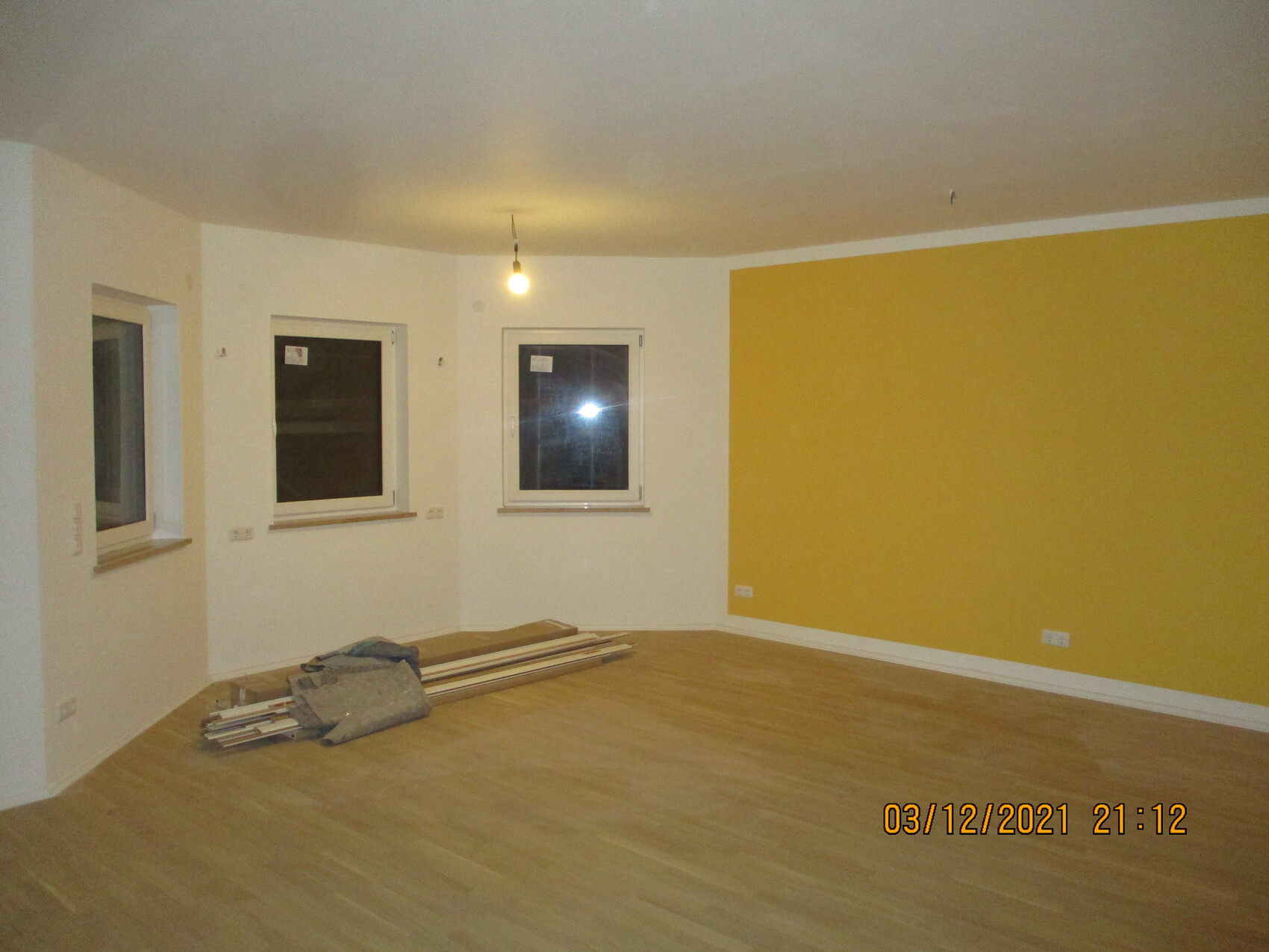 Leeres Zimmer: gelbe Wand, drei Fenster, Holzboden, Bauholzreste, hängende Glühbirne.