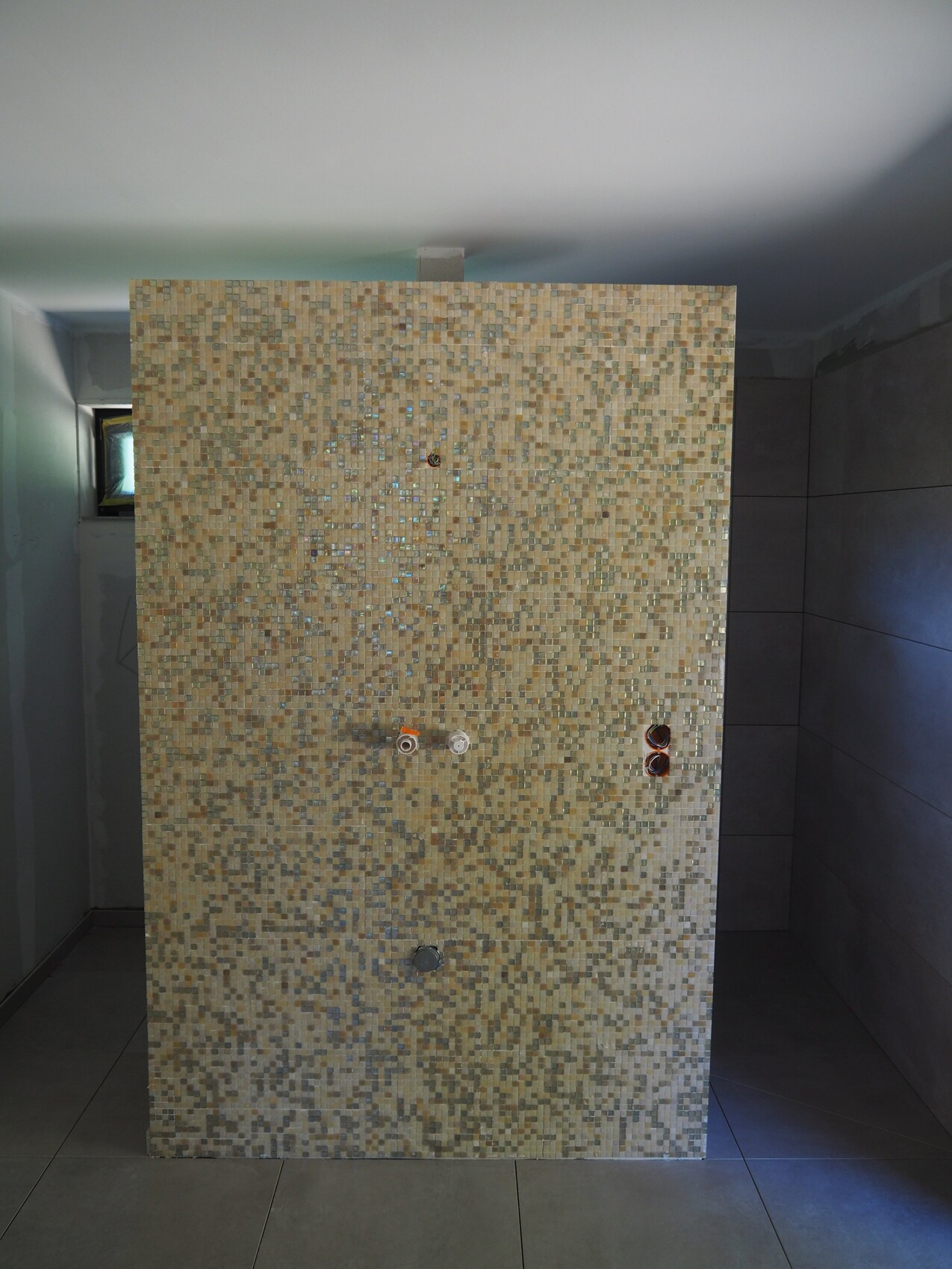 Beigefarbene Mosaikwand in Badezimmer/Dusche; sichtbare Wasserhähne, Rohranschlüsse und Abfluss.