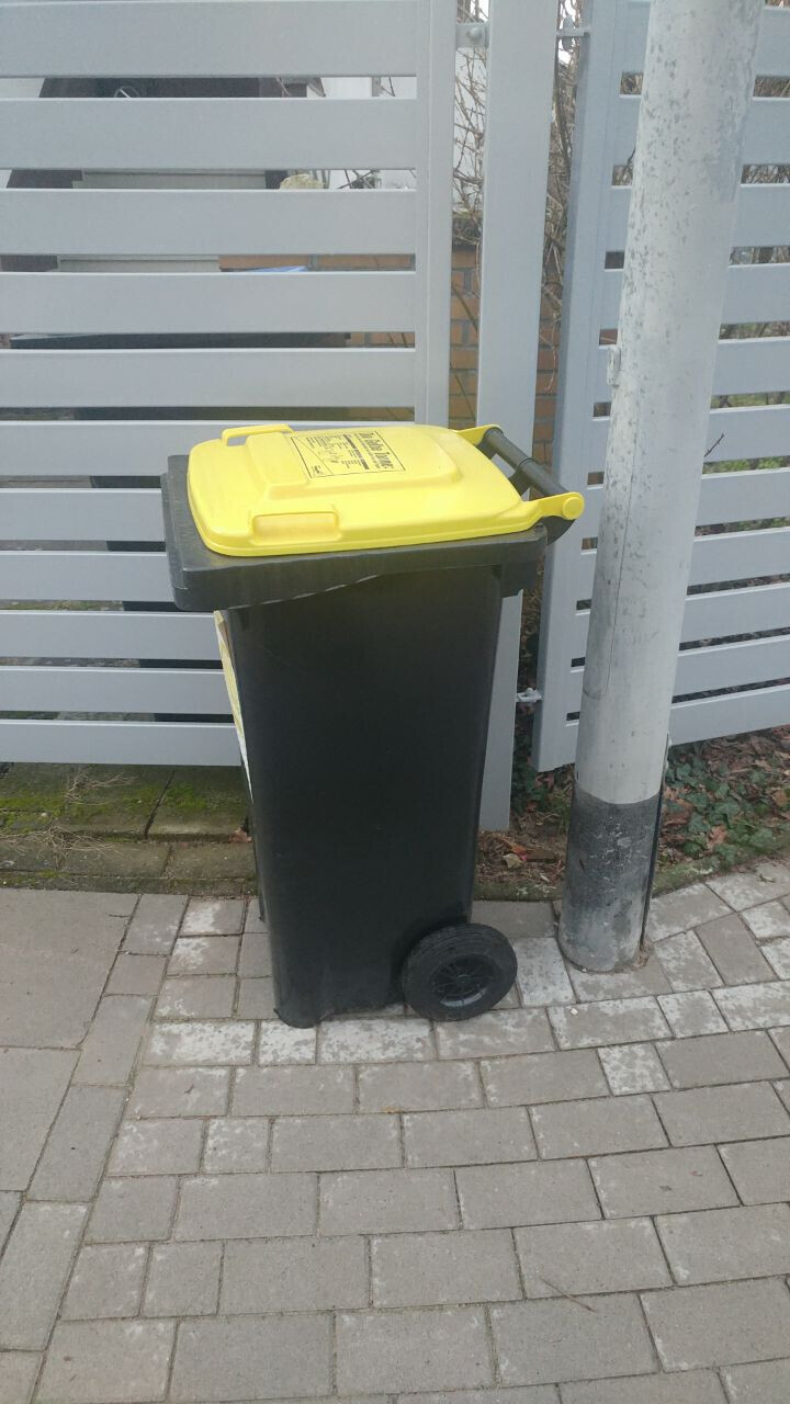 Schwarzer Abfallbehälter mit gelbem Deckel steht neben grauem Zaunpfosten auf dem Gehweg.