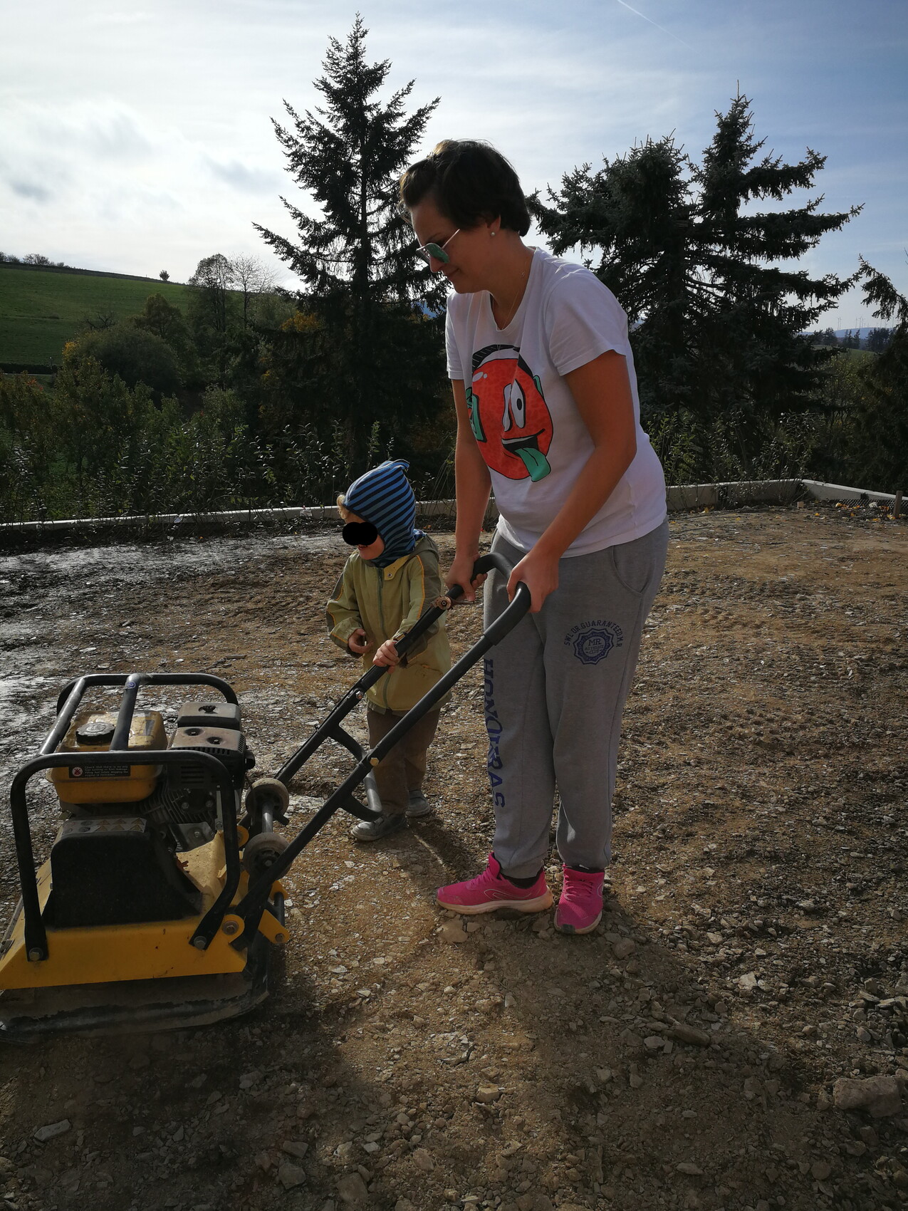 Frau mit Kind bedient eine Rüttelplatte auf einer Baustelle im Freien.
