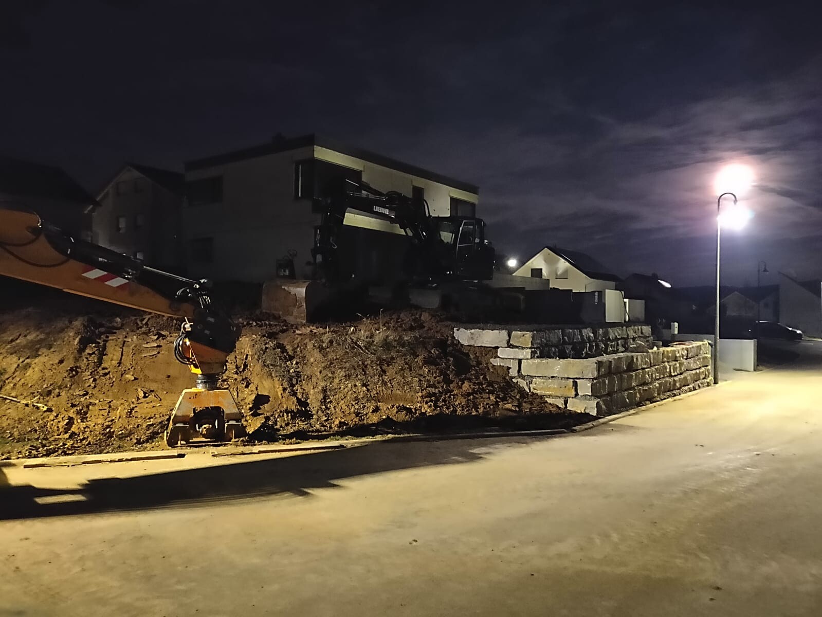Nächtliche Baustelle: Bagger auf Erdwall neben Steinmauer vor Häusern, Laterne leuchtet.